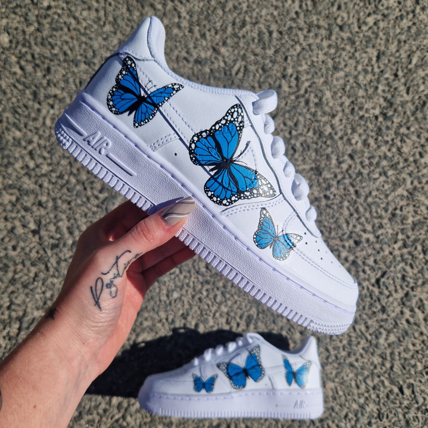 Nike Air Force 1 custom avec papillons bleus de tailles différentes peints à la main sur chaque chaussure.