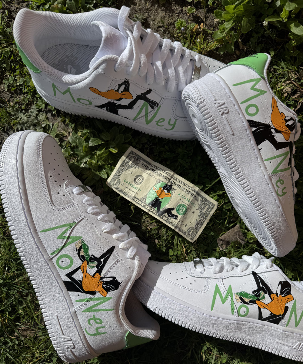Modèle présenté : Nike Air Force 1 Custom Daffy Duck Money, avec Daffy Duck peint différemment sur chaque chaussure et le mot "Money" en gros sur les côtés externes. Création réalisée à la main avec des peintures résistantes à l’eau, pour un rendu durable et original.