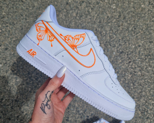 Nike Air Force 1 custom avec deux papillons orange fluo accrochés au swoosh externe entouré d’un halo lumineux orange