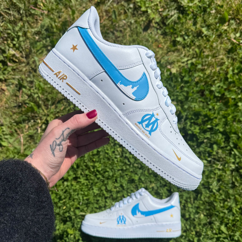 Nike Air Force 1 custom OM avec swoosh bleus et silhouette de la Bonne Mère, logo OM à l’avant, étoile dorée à l’arrière et swoosh internes en dégradé bleu et blanc.