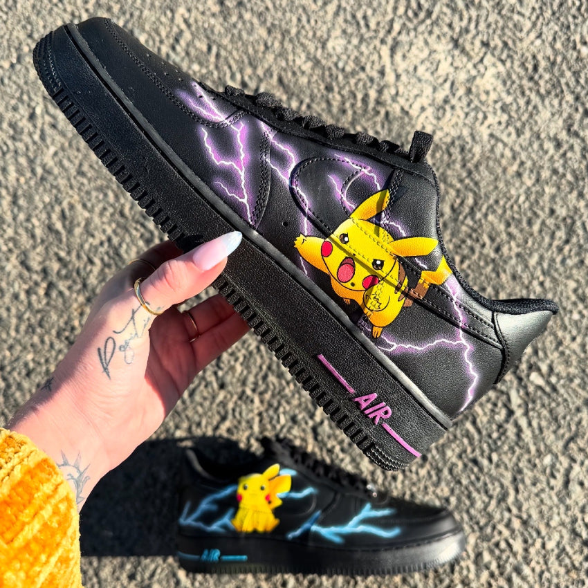 Nike Air Force 1 negras personalizadas con Pikachu en pleno ataque, rodeado de rayos violetas y azules en los laterales externos de cada zapatilla. Customización personalizable.