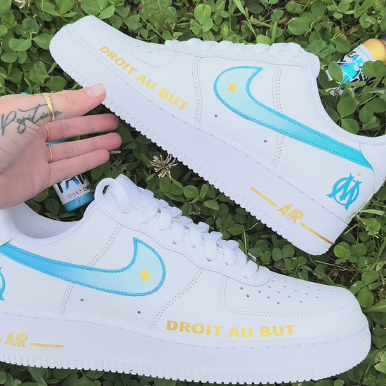 Modèle présenté : Nike Air Force 1 Custom OM – Droit au but. Swoosh dégradés bleu et blanc, étoile dorée, logo OM à l’arrière et inscription “DROIT AU BUT” en doré sur l’avant. Peinture à la main, finition résistante à l’eau, pour une paire unique et fidèle à l’esprit du club.